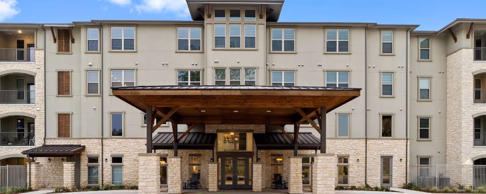 Front exterior view of a multi-story senior living facility with a covered entrance supported by stone pillars, large windows, and balconies on the sides under a partly cloudy sky.