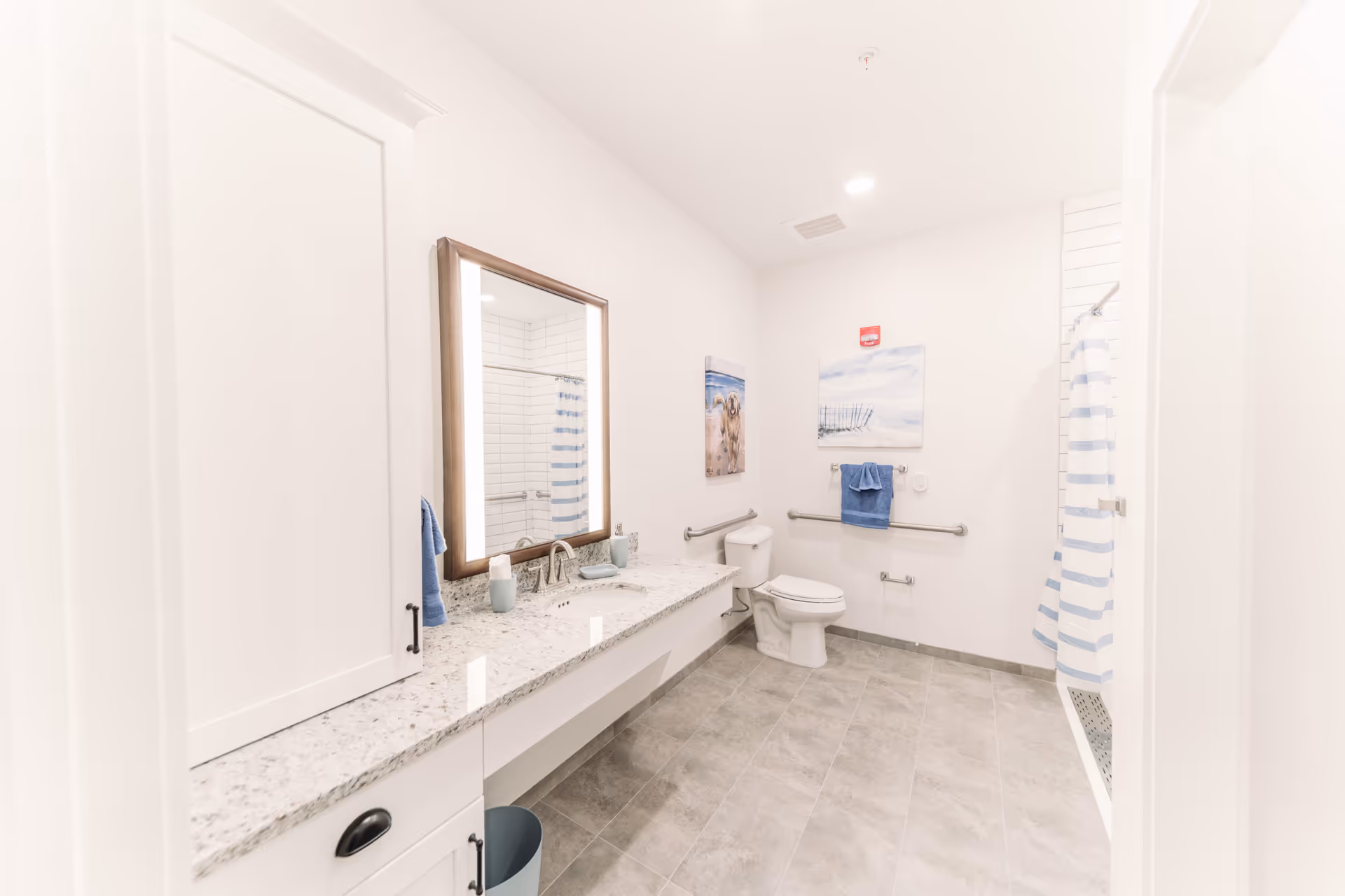 Bright and spacious bathroom with a granite countertop, a large mirror with wooden frame, a white toilet with grab bars on the walls, a shower with a blue and white striped curtain, blue towels, and two beach-themed paintings on the wall.