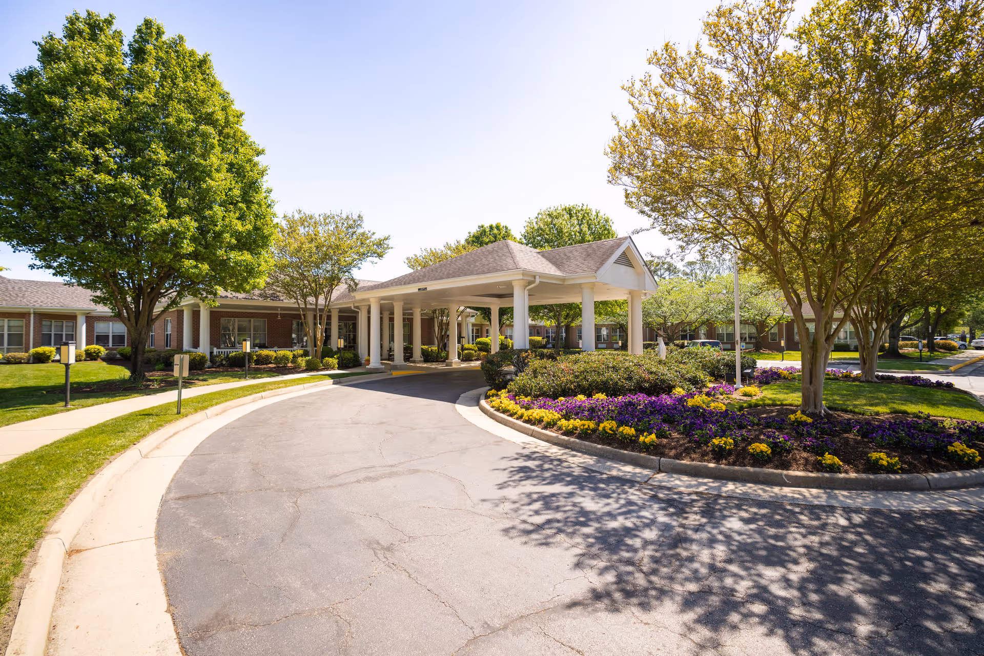 Exterior view of a senior living facility with a circular driveway, a covered entrance supported by white columns, well-maintained landscaping including green trees and colorful flower beds, and a clear blue sky.