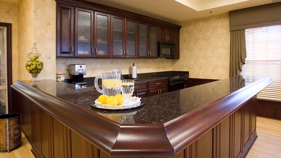 A kitchen area with dark wood cabinetry and a large granite countertop island. On the countertop, there is a tray with a pitcher of lemon water, a glass, and several lemons. A glass jar filled with green apples is placed on the left side. The kitchen has a microwave, stove, and a window with blinds and a valance.