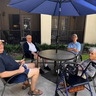 Four elderly individuals sitting around a round outdoor table with a large blue umbrella. They are seated on chairs on a concrete patio area with greenery and a building with windows in the background. One person has a walker nearby.