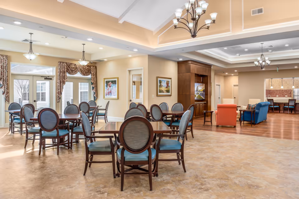 Spacious senior living common area with multiple dining tables and chairs, lounge seating, and chandeliers.