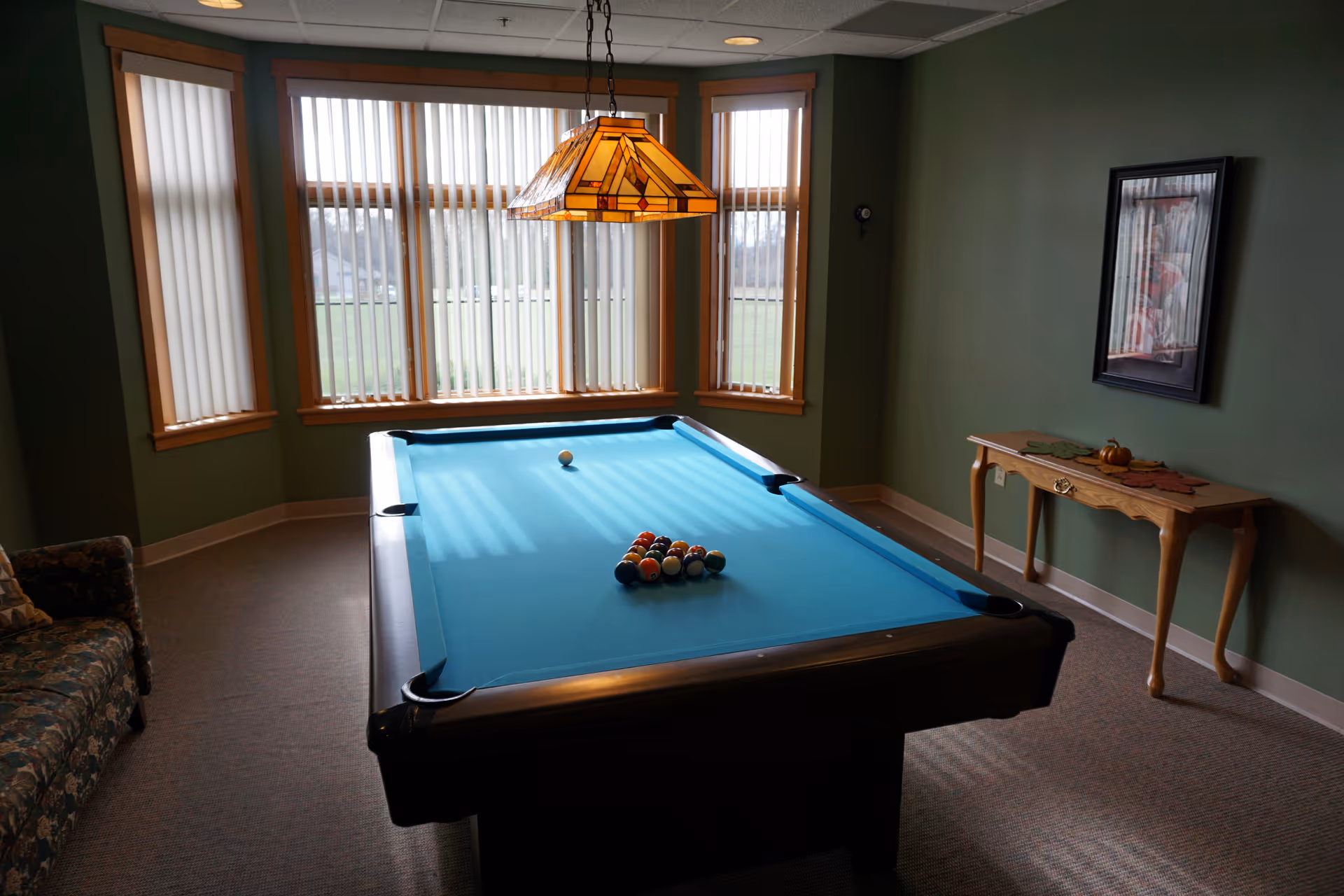 A room with a pool table set up for a game, featuring a blue felt surface and a rack of billiard balls. The room has green walls, a bay window with vertical blinds letting in natural light, a hanging stained glass lamp above the pool table, a floral patterned couch on the left, and a wooden side table with decorative items on the right wall.