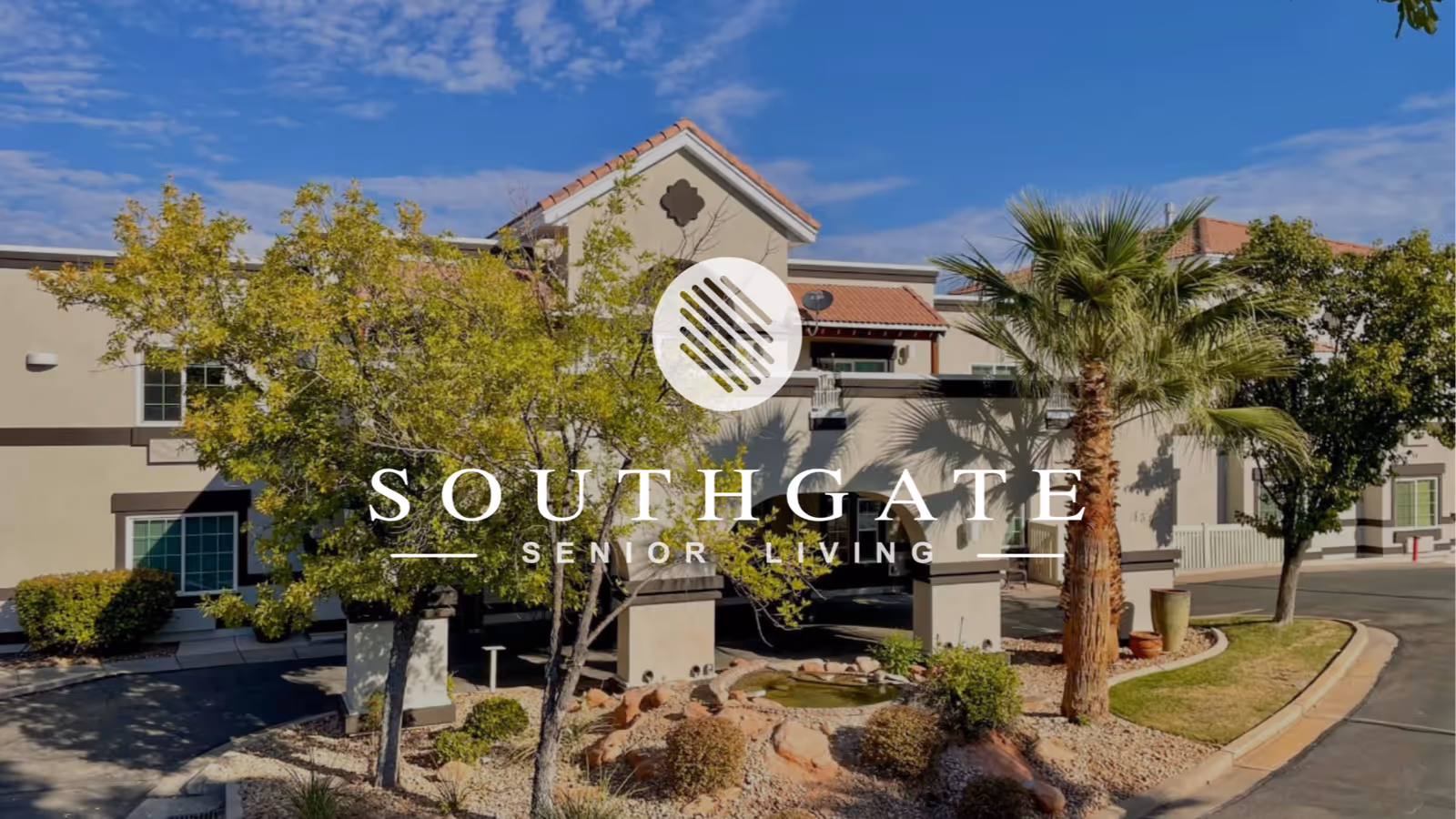 Exterior view of Southgate Senior Living facility showing a beige two-story building with a tiled roof, surrounded by trees and landscaping under a partly cloudy blue sky.