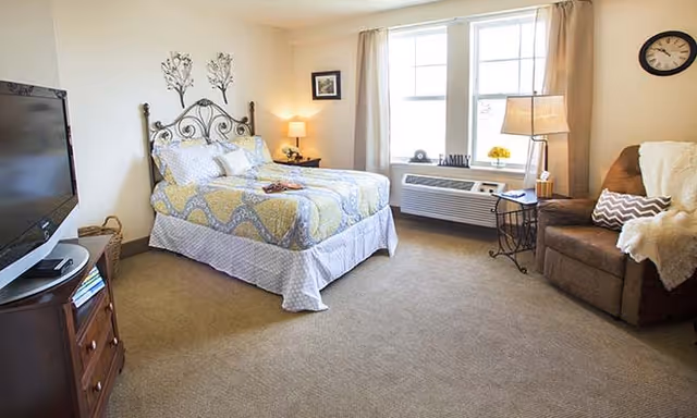 A cozy bedroom with a bed featuring a decorative metal headboard and patterned bedding. There is a wooden nightstand with a lamp and a small picture frame beside the bed. A comfortable armchair with a throw blanket and pillow is placed near a window with beige curtains. A wall clock hangs above the armchair. A flat-screen TV sits on a wooden dresser with shelves holding books and other items. The room is carpeted and well-lit with natural light coming through the window.