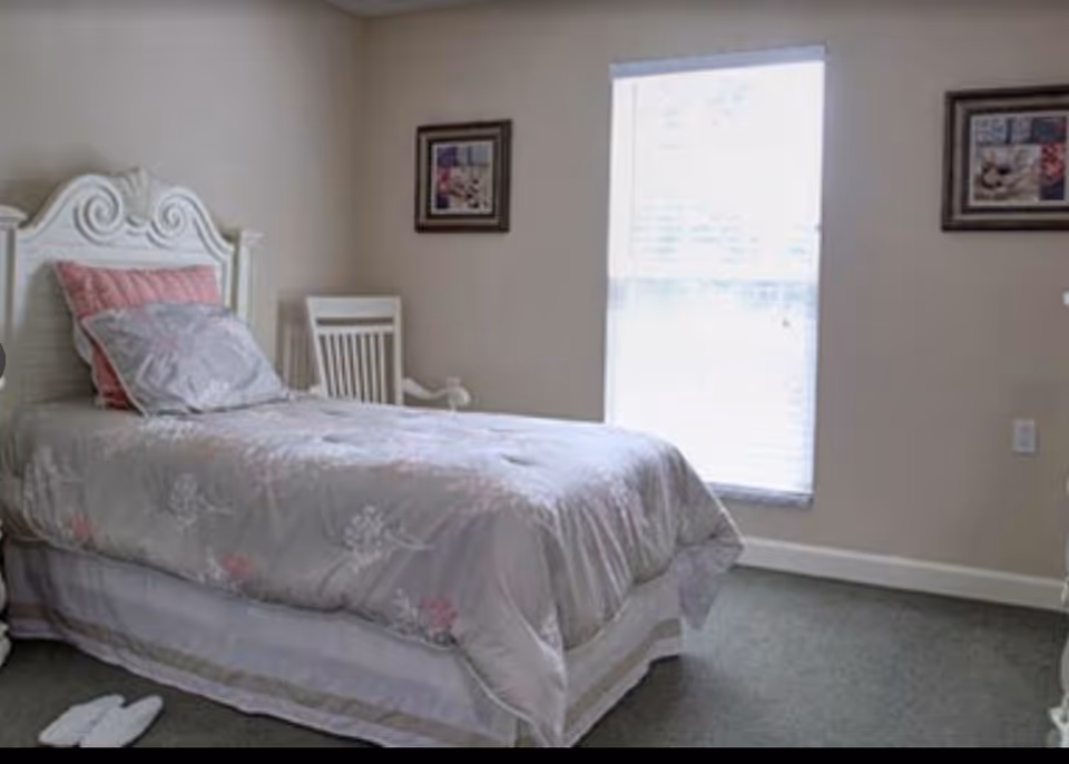 A bedroom with a single bed featuring a white ornate headboard, a gray and pink floral bedspread, and pillows. There is a white wooden chair next to the bed, two framed pictures on the beige walls, a window with blinds letting in natural light, and a pair of white slippers on the carpeted floor.