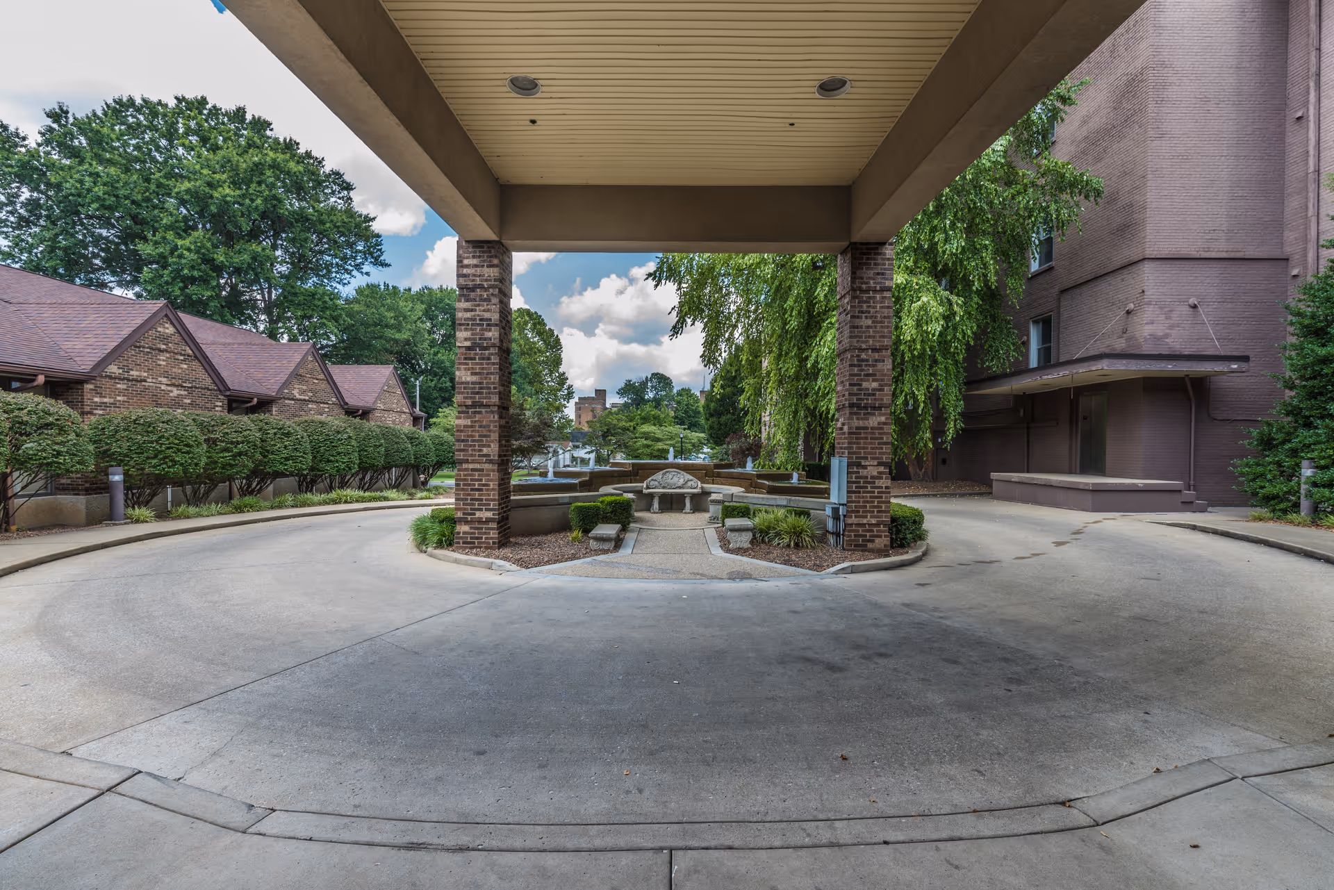Covered circular driveway and entrance (porte-cochere) of a senior living facility with brick columns, a central fountain and benches surrounded by landscaping.