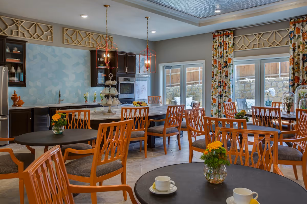 A bright and inviting dining area with multiple round tables and orange wooden chairs with gray cushions. The room features a kitchen area with dark cabinets, a light blue geometric backsplash, and stainless steel appliances. Two pendant lights hang over a marble island with a tiered tray of cups and a plate of fruit. Large windows with floral curtains let in natural light and show an outdoor stone wall and patio seating.