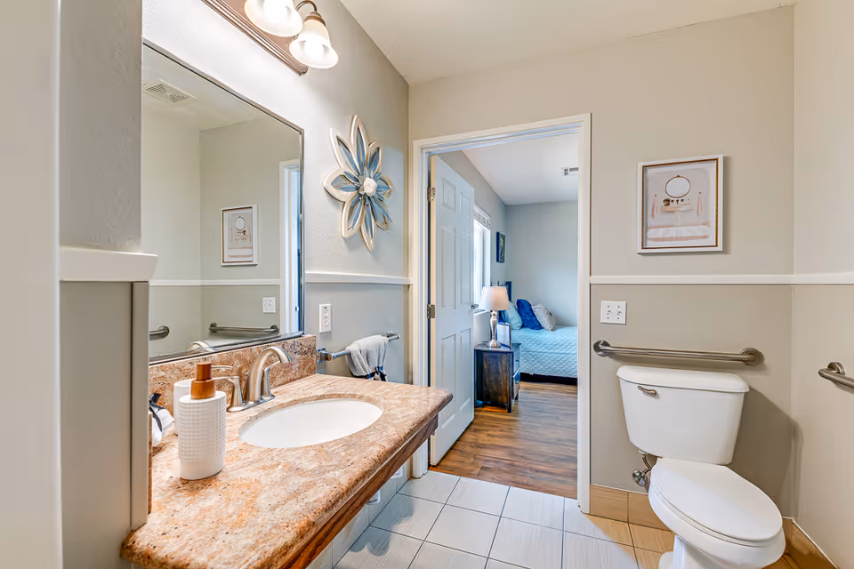 A bathroom with a granite countertop sink, a large mirror, and a toilet with grab bars on the walls. The bathroom has beige and white walls with a decorative flower wall art. Through an open door, a bedroom with a bed, nightstand, and lamp is visible.