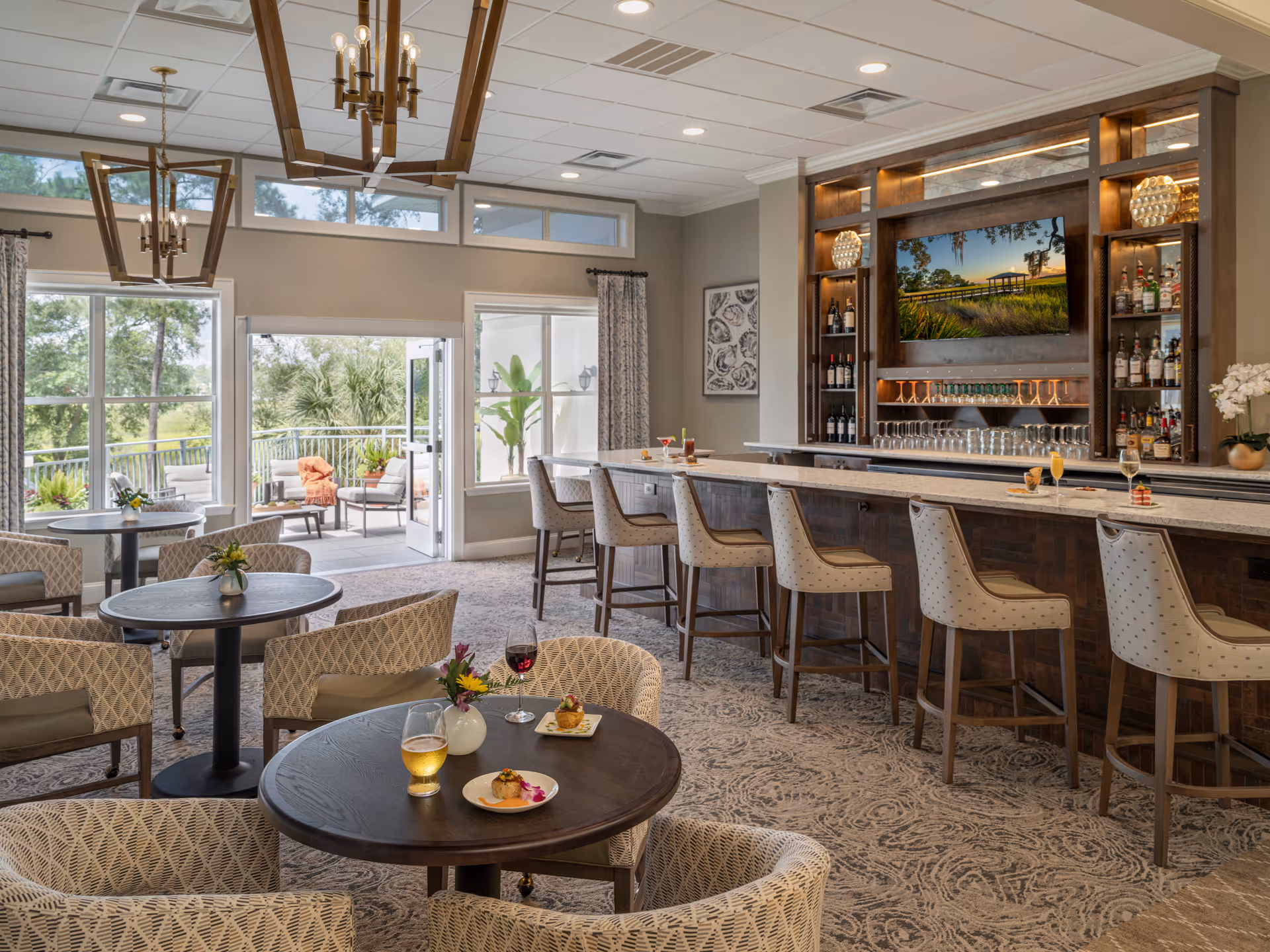 Bright lounge and bar area with round dining tables and upholstered chairs, a long marble-topped bar with stools, a wall-mounted TV, and open doors leading to a patio.