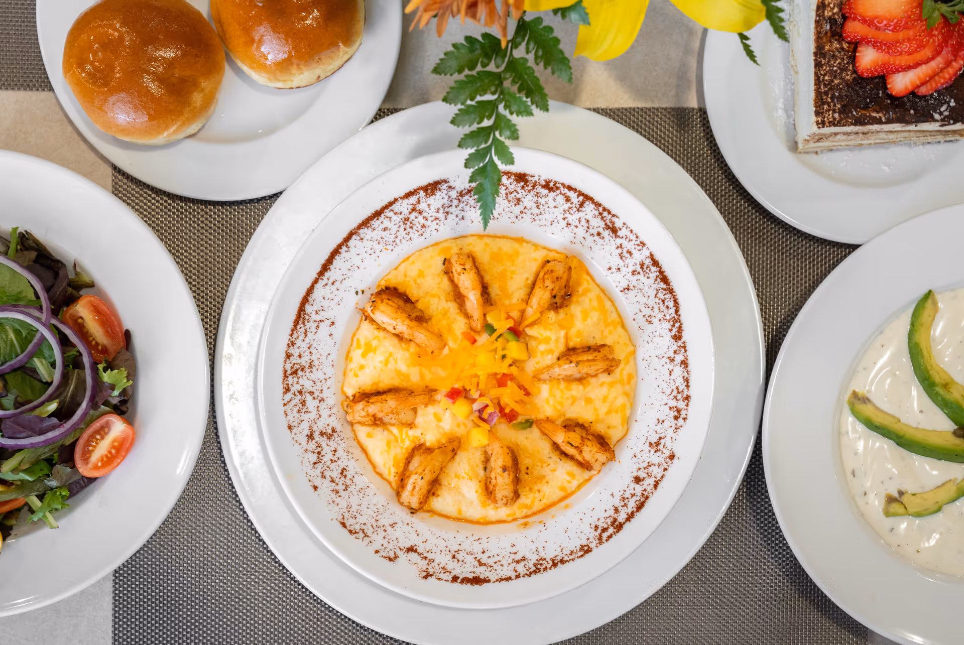 A top-down view of a meal consisting of a plate with shrimp arranged in a circle on a bed of cheesy grits, garnished with diced vegetables and a green leaf. Surrounding the main plate are other dishes including a salad with cherry tomatoes and red onion, two bread rolls on a small plate, a dessert topped with sliced strawberries, and a plate with avocado slices on a creamy sauce.