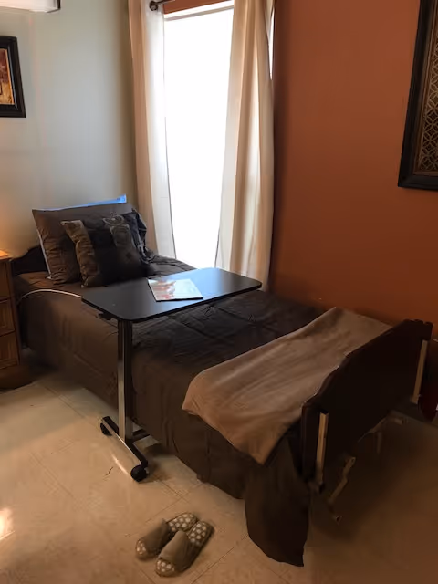 A single bed with brown bedding and multiple pillows is positioned next to a window with beige curtains. A small overbed table with a magazine on it is placed over the bed. A pair of slippers is on the floor near the foot of the bed. The room has a warm-toned wall and a framed picture hanging on it.