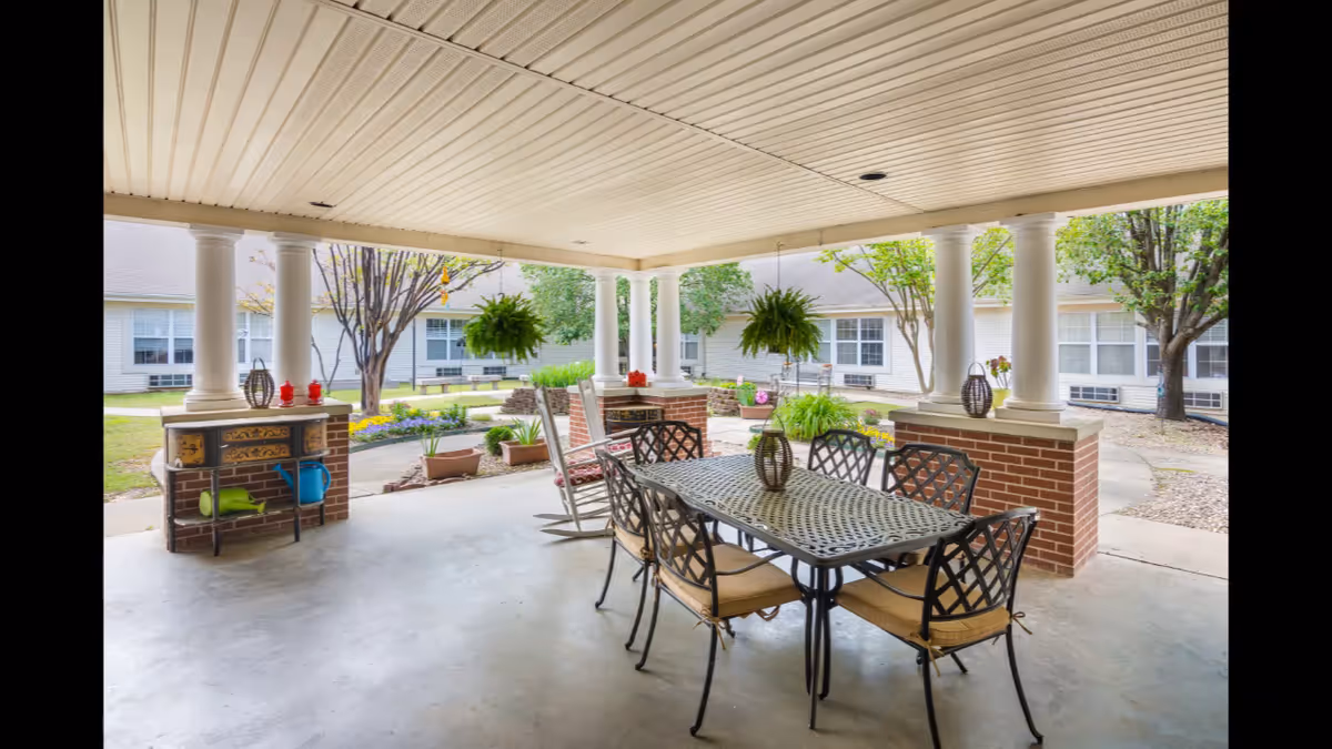 Covered outdoor patio area with a metal table and six chairs with cushions, two rocking chairs, hanging plants, and decorative items on brick pillars. The patio overlooks a garden with trees, flowers, and a walkway, surrounded by a building with multiple windows.