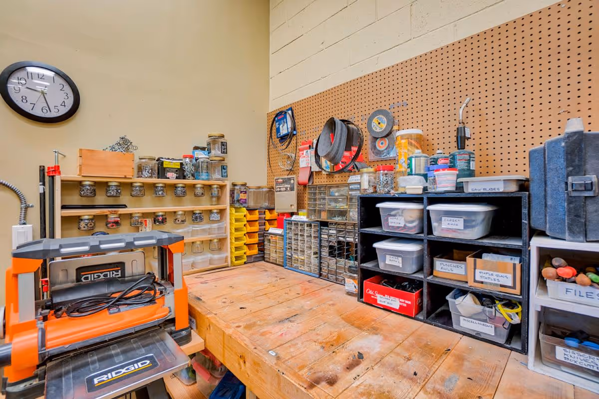 A well-organized workshop area with a wooden workbench, various small storage containers filled with screws and hardware, a pegboard on the wall holding tools and supplies, and a Ridgid power tool on the left side. A clock is mounted on the beige wall above the workbench.