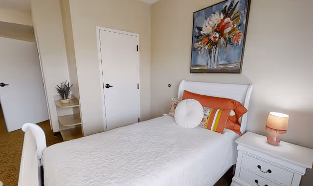 A neatly made single bed with white bedding and colorful pillows in a small bedroom. There is a white nightstand with a pink lamp on the right side of the bed. A floral painting hangs on the wall above the bed. Two closed white doors and a small corner shelf with a plant are visible in the background.