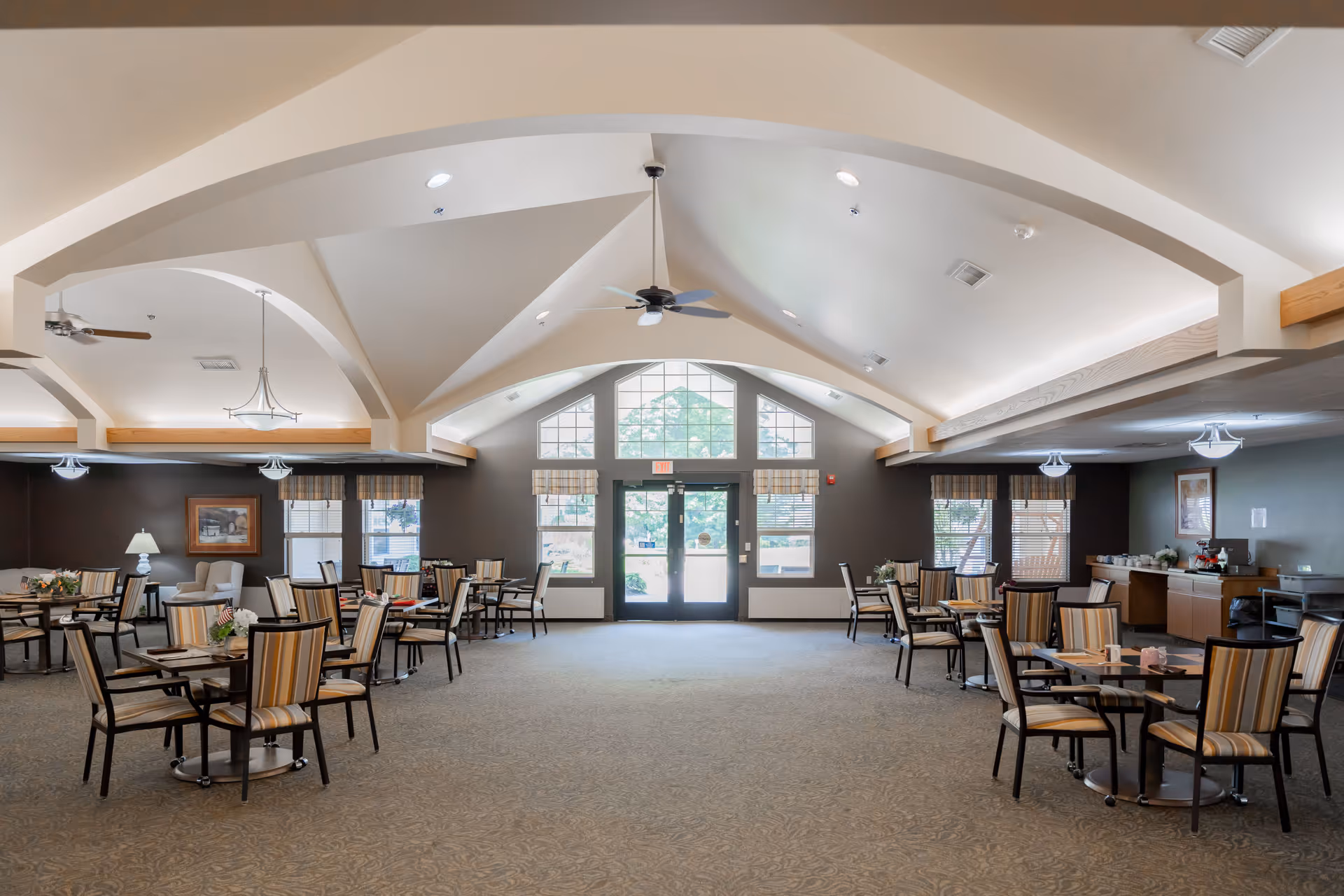 Interior view of the dining room at Tawas Village, featuring tables with striped chairs, large windows, and a high ceiling with decorative lighting.