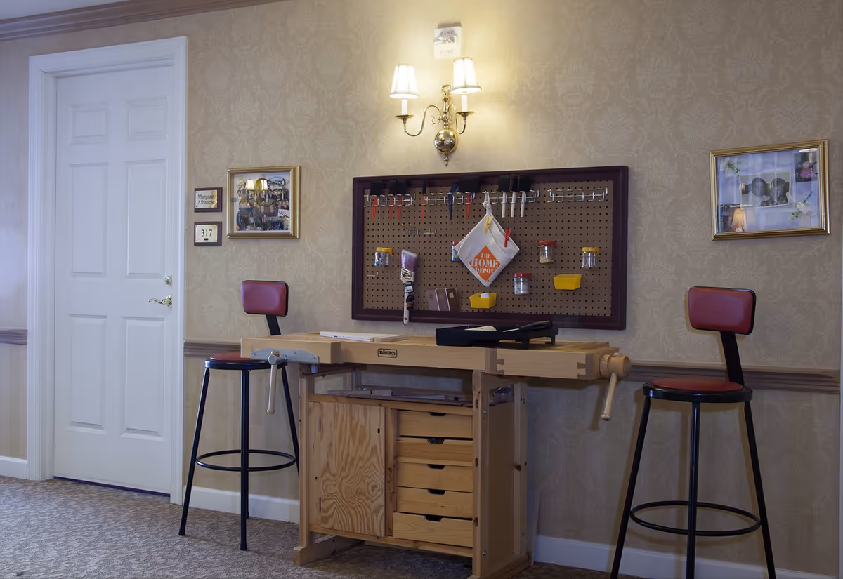 Wooden craft workbench with tools on a pegboard and two red-cushioned stools against a patterned wallpapered wall in an interior room.