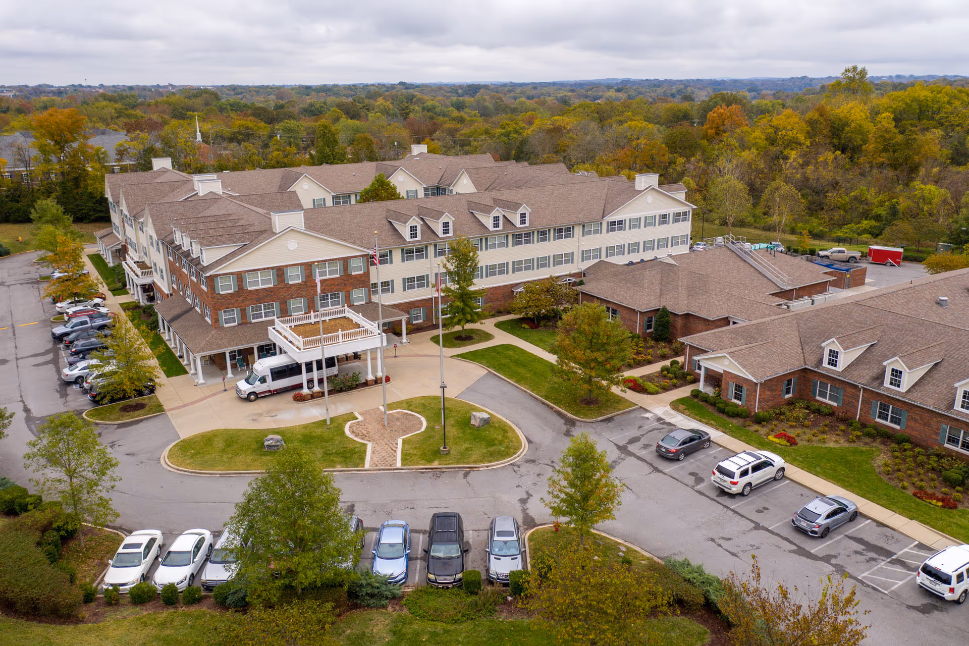 Aerial view of Vitality Living Hendersonville, a large senior living facility with multiple connected buildings featuring brick and beige siding exteriors, surrounded by trees with autumn foliage. The facility has a circular driveway with flagpoles and several parked cars in the parking lot.