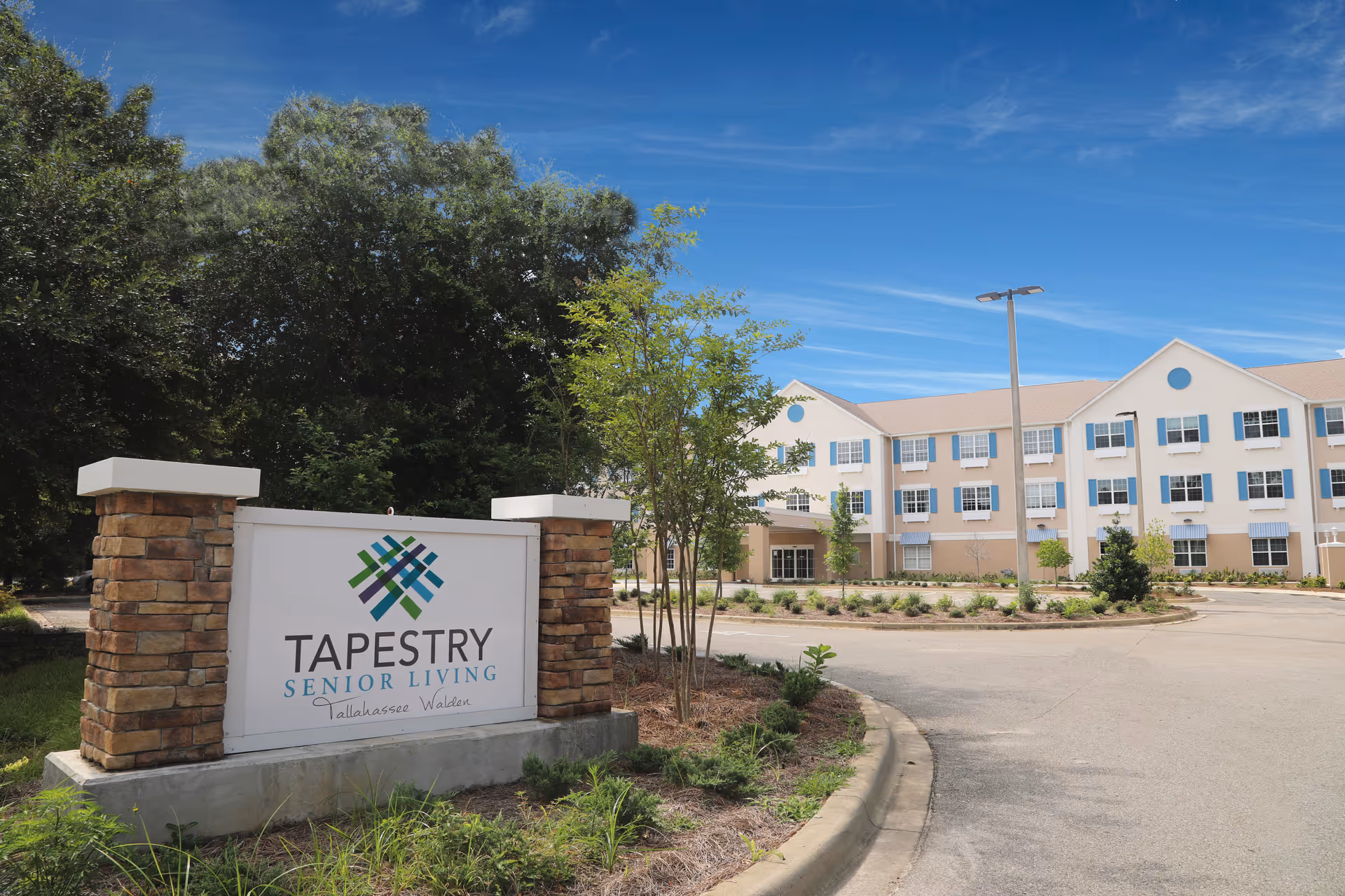 Exterior view of Tapestry Senior Living Walden facility with a large sign in the foreground displaying the facility name. The building is a three-story structure with multiple windows, beige and white walls, and a clear blue sky above. There are trees and landscaped greenery around the sign and building.
