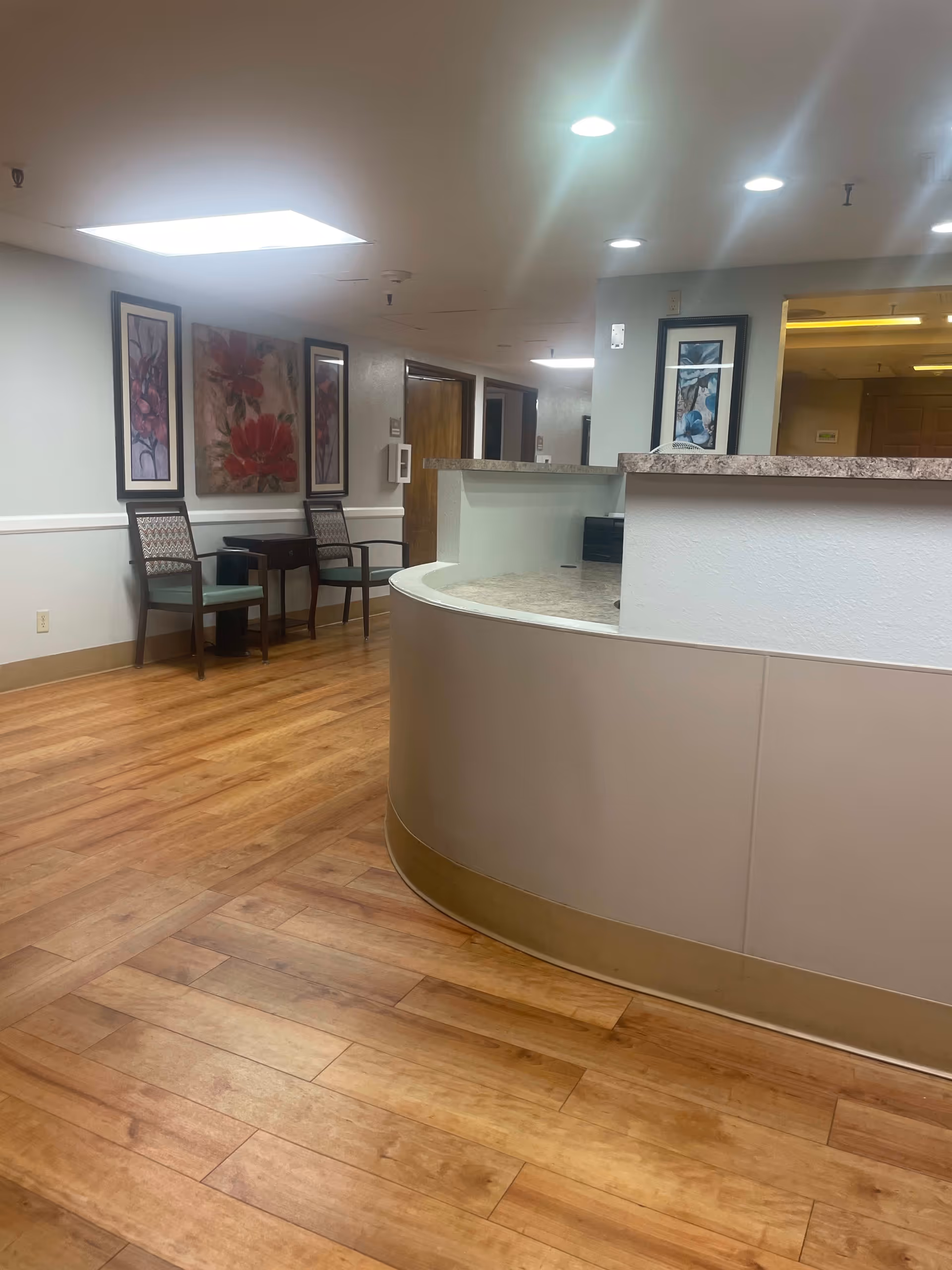 Interior view of a senior living facility hallway with a curved reception desk on the right, wooden flooring, two chairs and a small table against the left wall, and framed floral artwork hanging above the chairs.