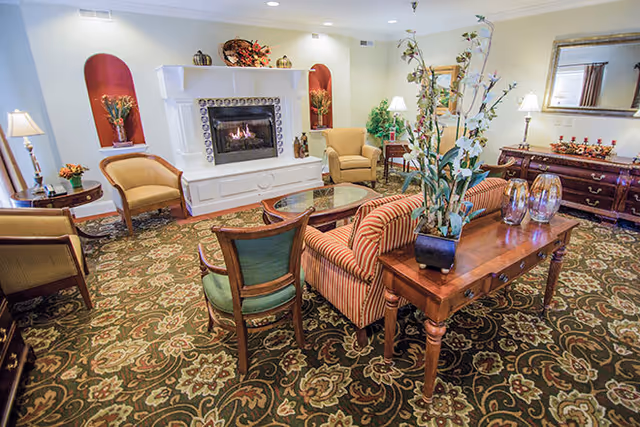 A cozy living room with a patterned carpet, a white fireplace with a fire burning, several upholstered chairs and a striped sofa around a glass coffee table. There is a wooden side table with a potted plant and decorative glass items, a large mirror on the wall, and soft lighting from lamps.