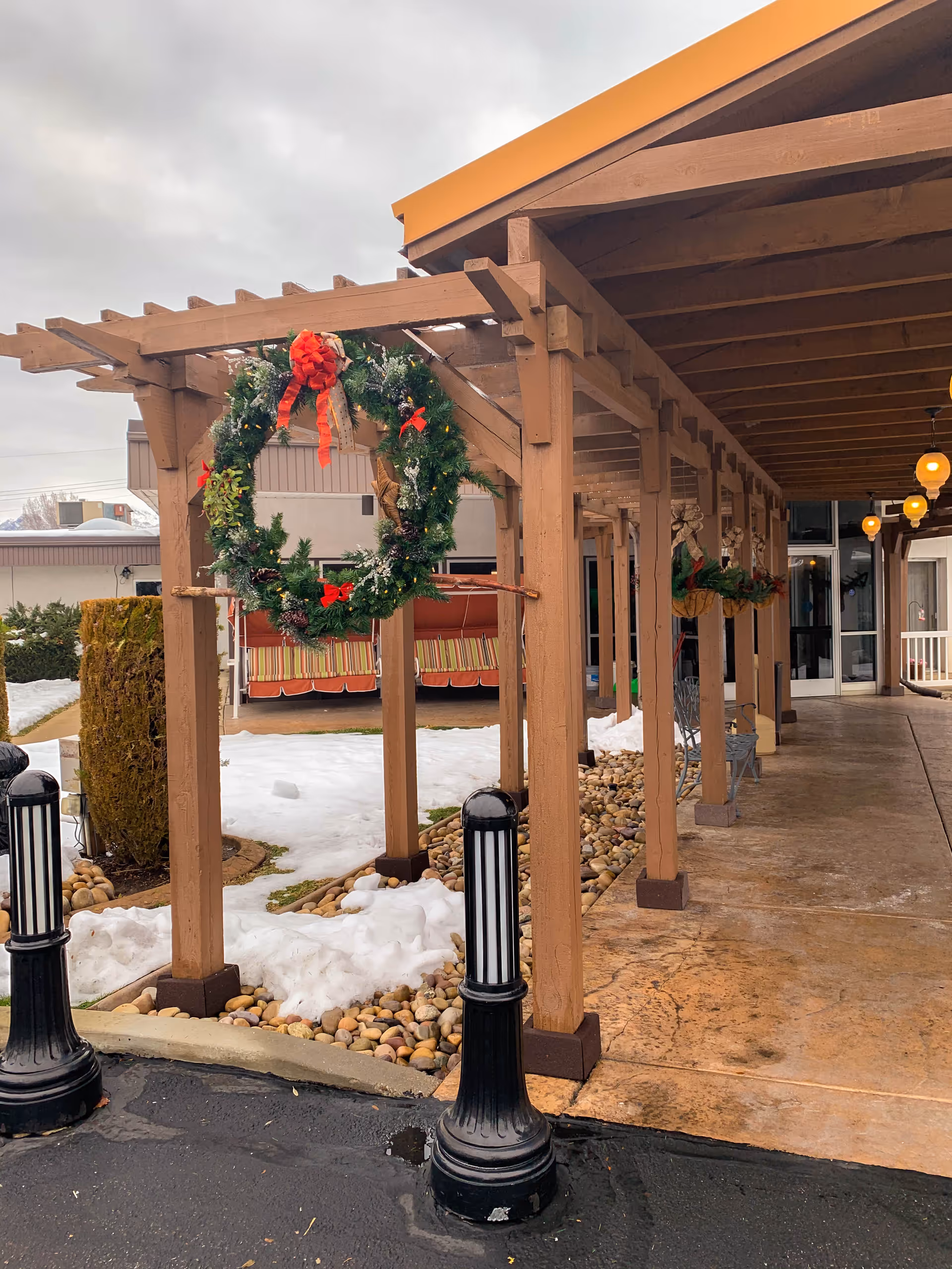 Outdoor covered walkway with wooden beams decorated with Christmas wreaths. Snow is on the ground beside the walkway, and there are black bollards along the edge of the pavement. The entrance to a building is visible at the end of the walkway.