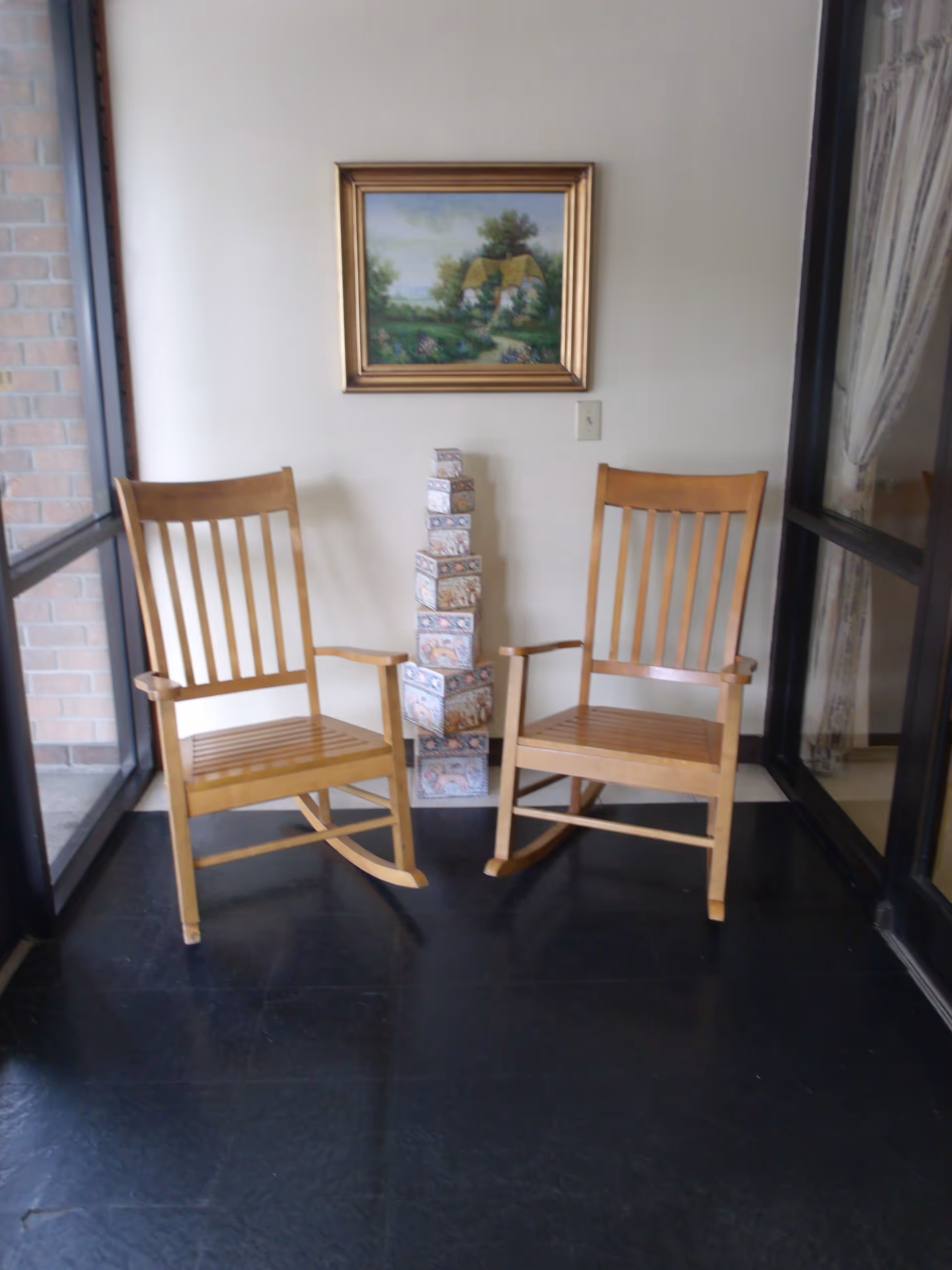 Two wooden rocking chairs placed side by side on a black floor in a corner with glass windows on both sides. Between the chairs is a decorative stack of patterned boxes. A framed painting of a countryside scene with a house and trees hangs on the wall above the boxes.