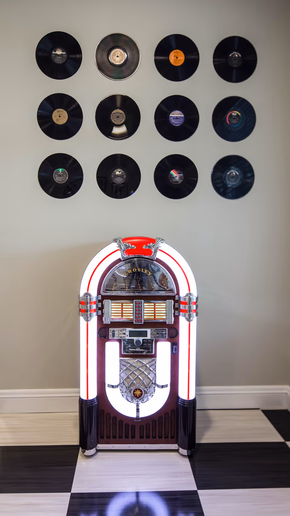 A vintage Crosley jukebox illuminated with white and red lights stands on a black and white checkered floor. Above it, twelve vinyl records are arranged in a grid pattern on a light-colored wall.