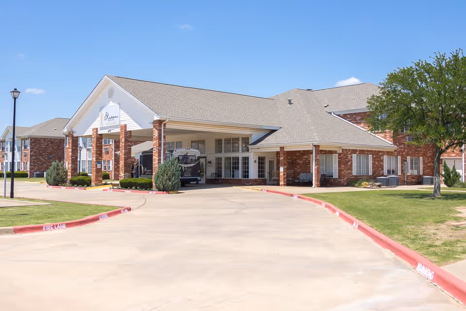 Exterior view of Elison Assisted Living of Lake Wellington showing a brick building with a covered entrance, a driveway, and a tree on the right side under a clear blue sky.