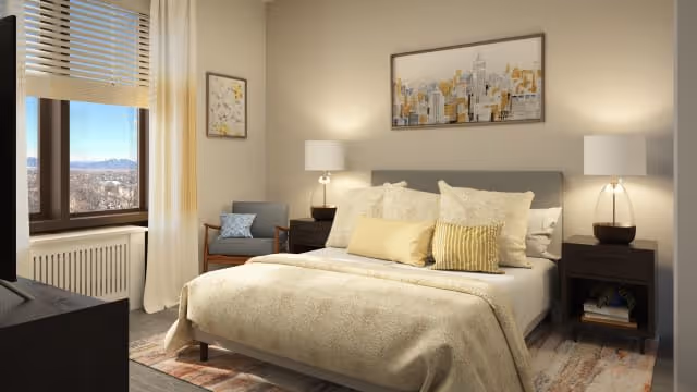 A cozy bedroom with a large bed covered in beige bedding and multiple pillows. There are two bedside tables with lamps on each side of the bed. A framed cityscape artwork hangs above the headboard. A window with blinds and curtains lets in natural light, and a comfortable chair with a cushion is placed near the window. The room has a warm and inviting atmosphere.