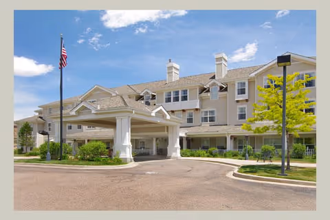 Exterior view of a multi-story senior living facility with beige siding, white trim, and a covered entrance. There is a flagpole with an American flag and some greenery around the building under a partly cloudy blue sky.