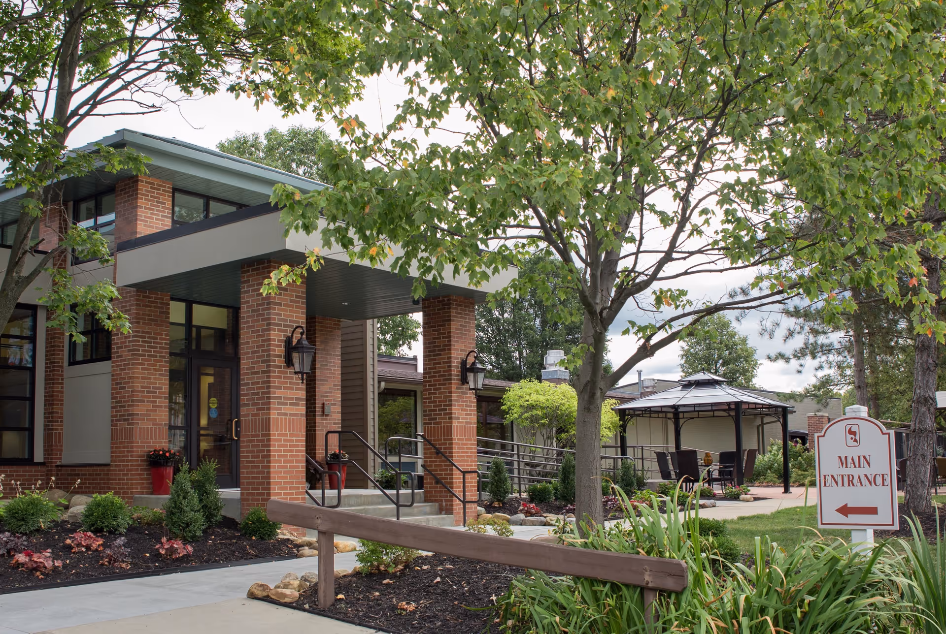 Exterior view of Cardinal Retirement Village showing the main entrance with brick pillars, a covered entryway, landscaped garden with shrubs and trees, a pathway leading to a gazebo with outdoor seating, and a sign indicating the main entrance.
