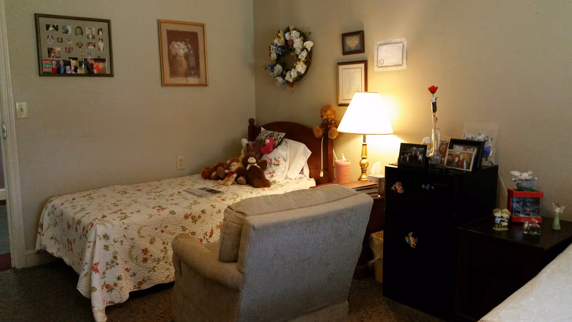 A cozy bedroom in a senior living facility with a single bed covered in a floral quilt, several stuffed animals on the bed, a cushioned armchair in front of the bed, a nightstand with a lamp, framed pictures and certificates on the wall, and a small black refrigerator with decorative magnets.