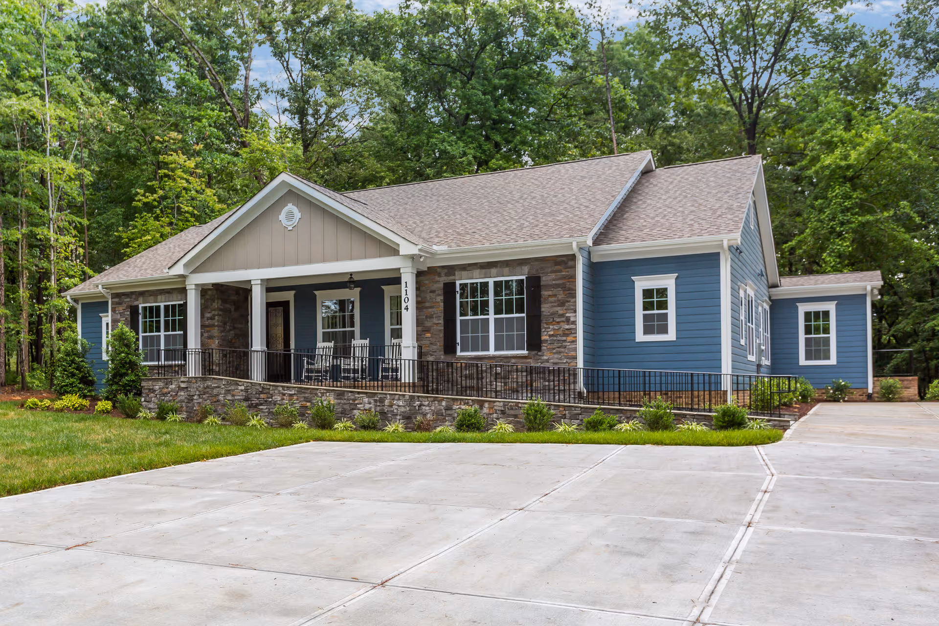 Exterior view of a single-story assisted living facility building with blue siding, stone accents, a covered porch with rocking chairs, surrounded by greenery and trees, and a concrete driveway in front.