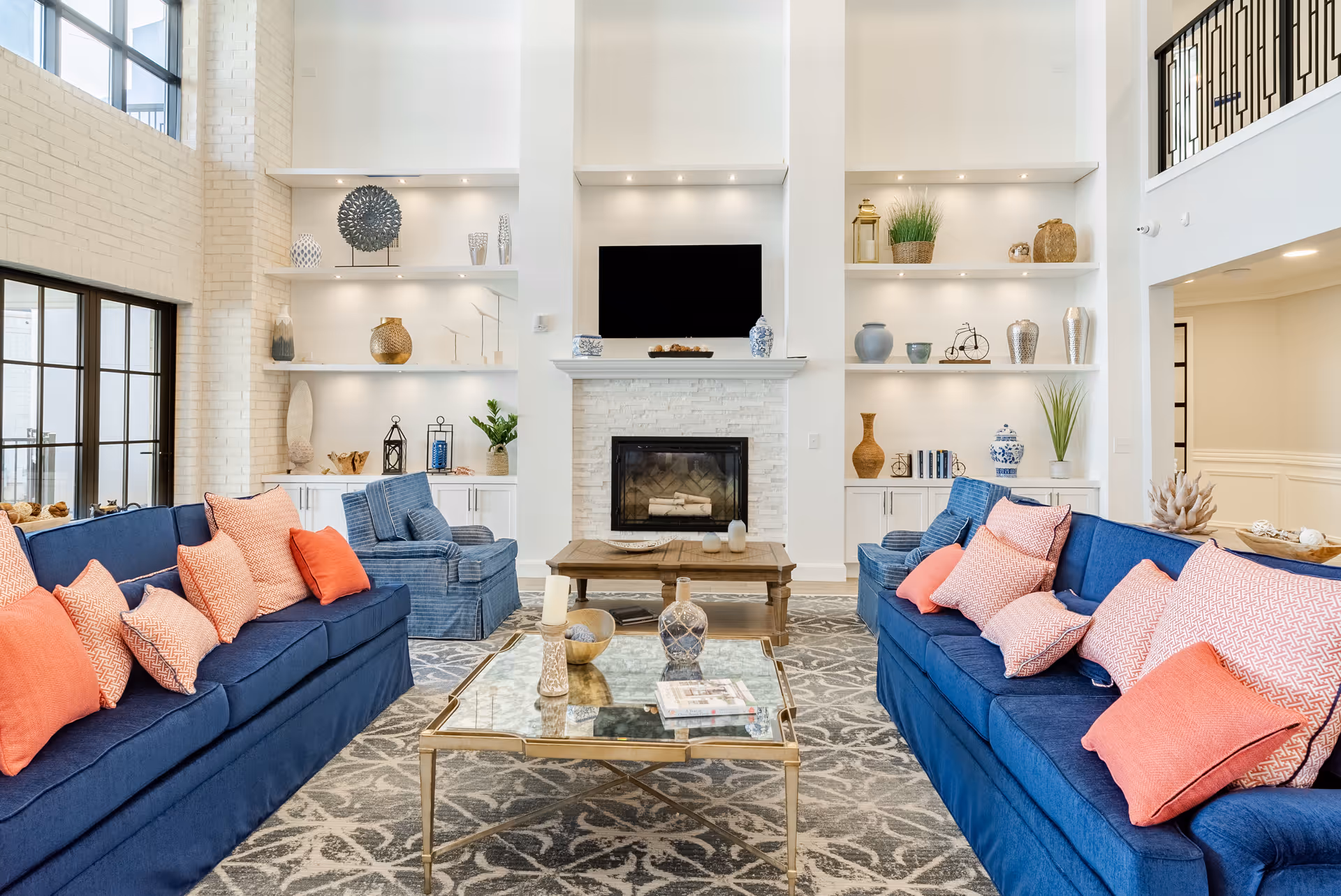 Bright, spacious living room with two blue sofas facing a fireplace and built-in shelving, accented by coral pillows and a glass coffee table.