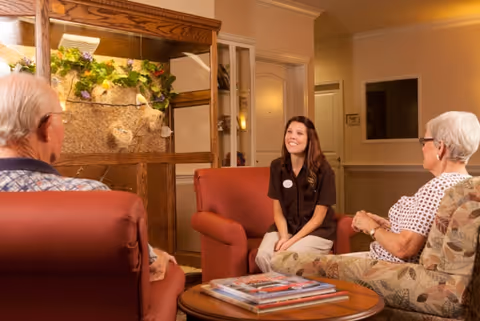 A young woman sitting and smiling while talking to two elderly people seated in a cozy living room area with comfortable chairs and a wooden display cabinet with decorative plants in the background.