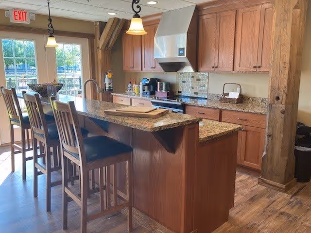 A bright kitchen area with wooden cabinets, granite countertops, and a kitchen island with four high chairs. The kitchen features a stove with a stainless steel hood, a coffee maker, and various kitchen items on the counter. There are pendant lights hanging from the ceiling and a glass door leading outside with an exit sign above it.