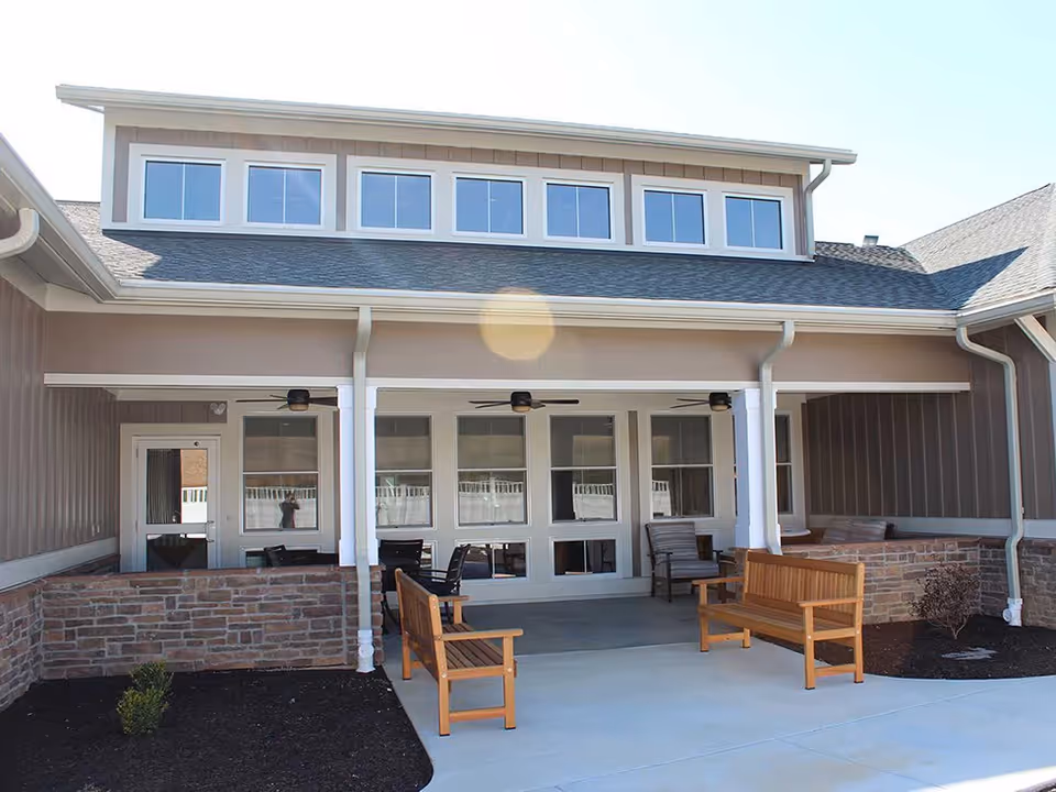 Covered front patio/entrance of a senior living building with wooden benches, chairs, ceiling fans, and large windows.