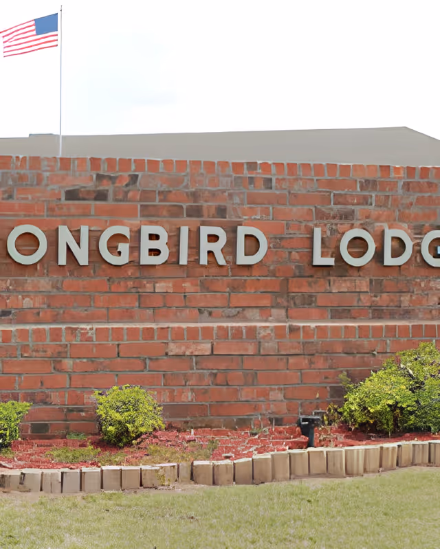 Brick entrance wall with metal letters spelling 'SONGBIRD LODGE', a small landscaped bed, and an American flag on a pole behind it.