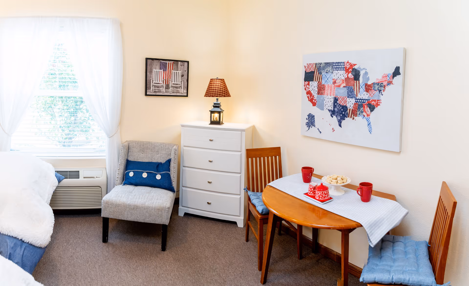 A cozy senior living room corner with a small wooden dining table set for two with red mugs and a plate of cookies, two wooden chairs with blue cushions, a gray upholstered chair with a blue pillow, a white chest of drawers with a lamp on top, and wall art including a map of the United States and a picture of rocking chairs with an American flag. A window with sheer white curtains lets in natural light.