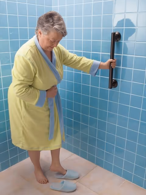 An older woman in a robe steadies herself on a grab bar inside a blue-tiled shower with slippers on the floor.