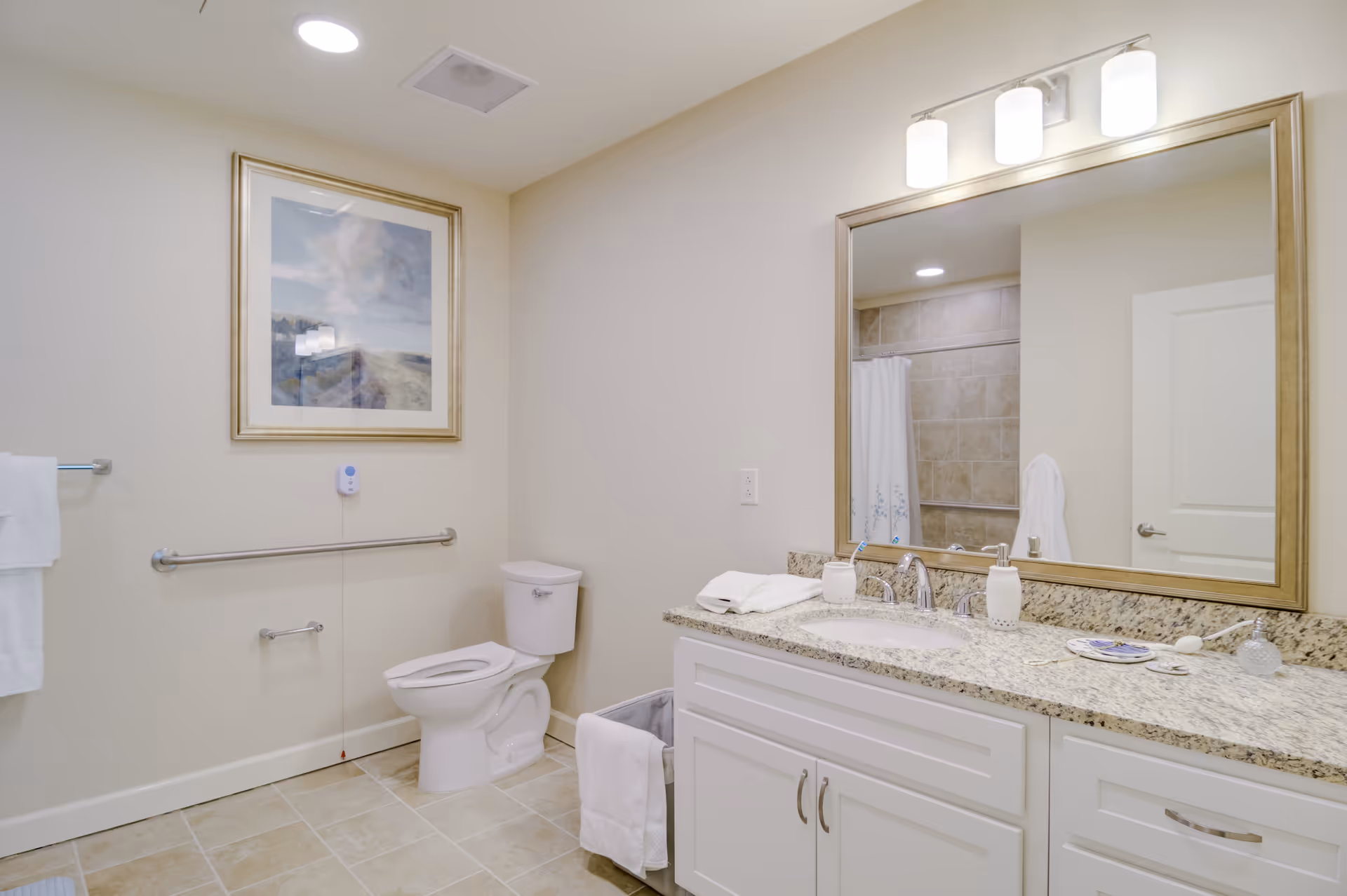 A clean and well-lit bathroom with beige tiled floor and walls painted in a light cream color. The bathroom features a white toilet with a grab bar on the wall beside it, a large framed mirror above a granite countertop with a white sink, and white cabinetry below. There is a towel rack with white towels on the left wall, a framed landscape picture above the grab bar, and a shower with a white curtain visible in the mirror's reflection. The lighting fixture above the mirror has three white cylindrical lights.