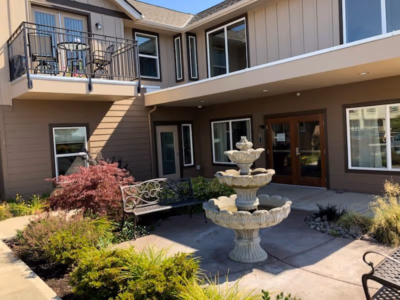 Outdoor courtyard area of McMinnville Senior Living Apartments featuring a three-tiered stone fountain, metal benches, landscaped plants, and the exterior of the building with windows and a balcony.