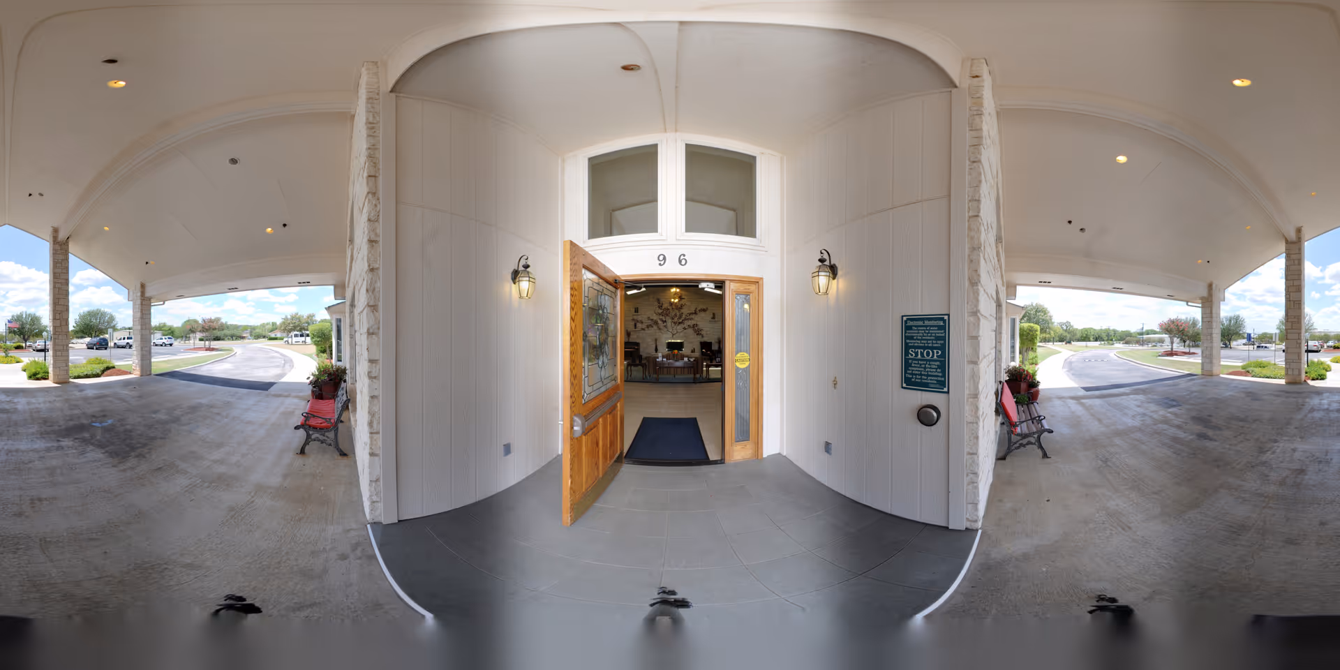 Entrance to Heritage Place at Fredericksburg showing an open wooden door with glass panels leading into a lobby area. The entrance is covered by a large canopy supported by stone pillars. There are benches on either side of the entrance and a driveway curving around the building. The sky is clear with some clouds.