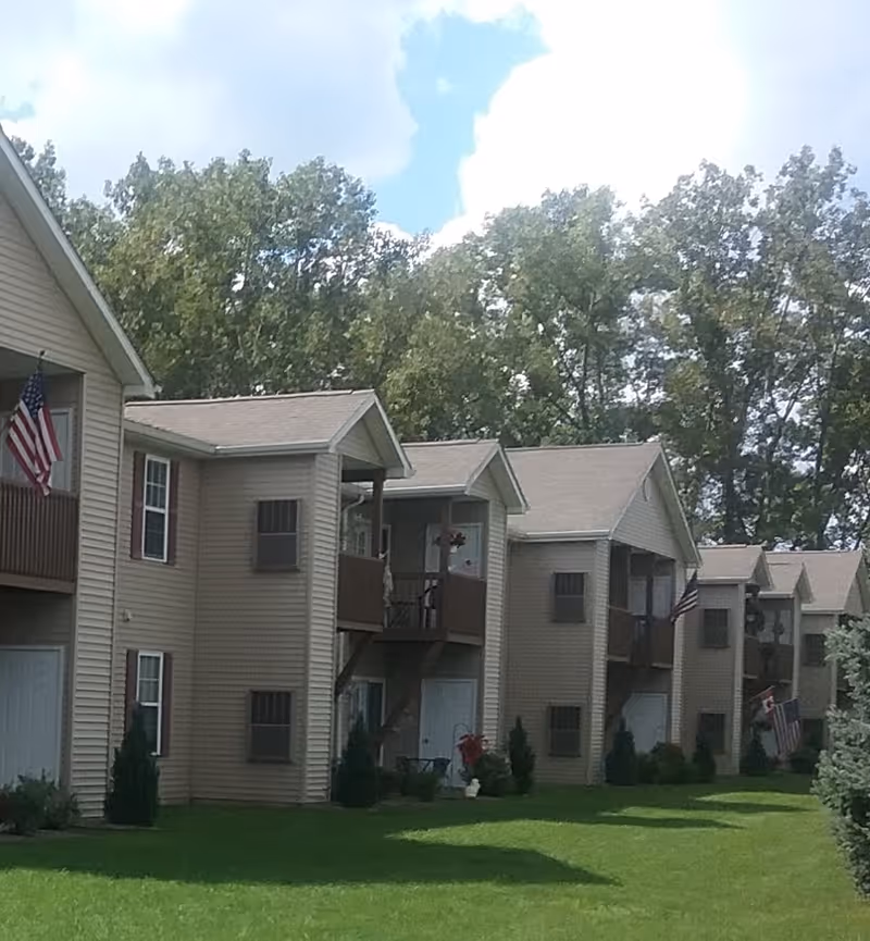 Exterior view of a row of beige two-story senior apartment buildings with balconies, green lawn, and trees in the background under a partly cloudy sky.