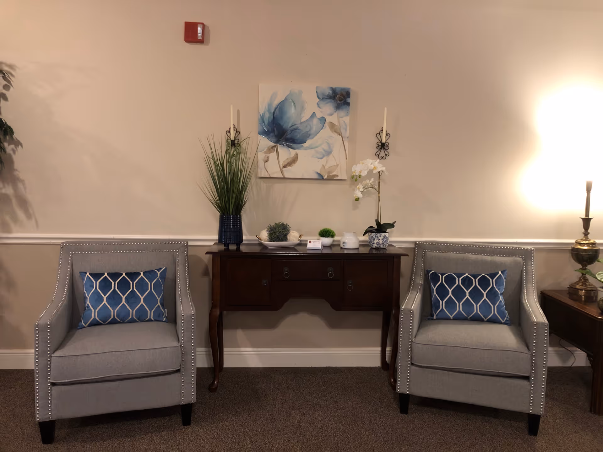 A cozy interior seating area with two gray armchairs featuring blue patterned cushions. Between the chairs is a dark wooden console table decorated with various plants and small decorative items. Above the table hangs a painting of blue flowers, flanked by two wall-mounted candle holders. A lamp on a side table to the right provides warm lighting.
