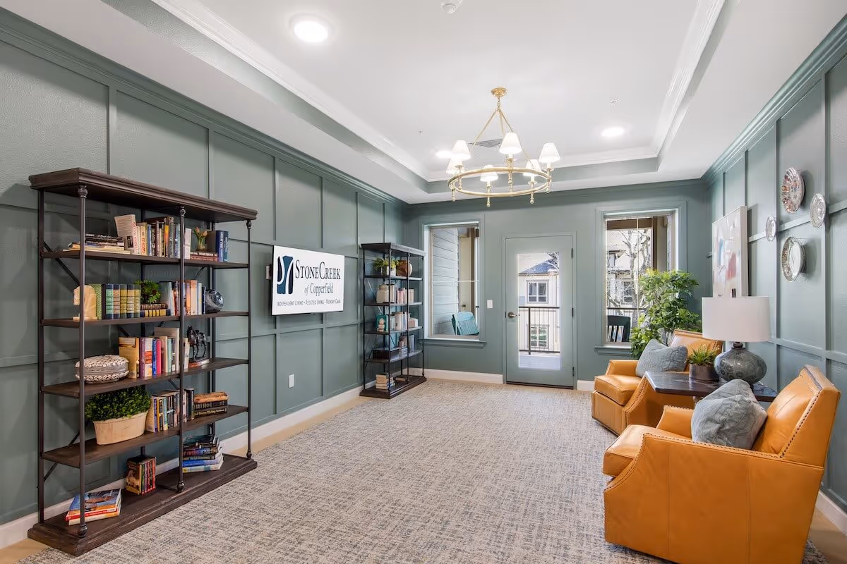 A cozy senior living common area with green paneled walls, two mustard yellow armchairs with gray cushions, a side table with a lamp and plant, two bookshelves filled with books and decor, a wall-mounted TV displaying the StoneCreek of Copperfield logo, and a door with windows leading outside.