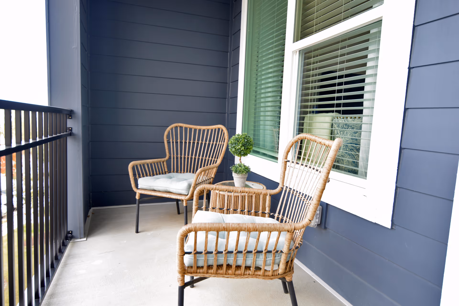 A small balcony with two wicker chairs, a round side table holding a potted plant, a metal railing, and a window set into blue siding.