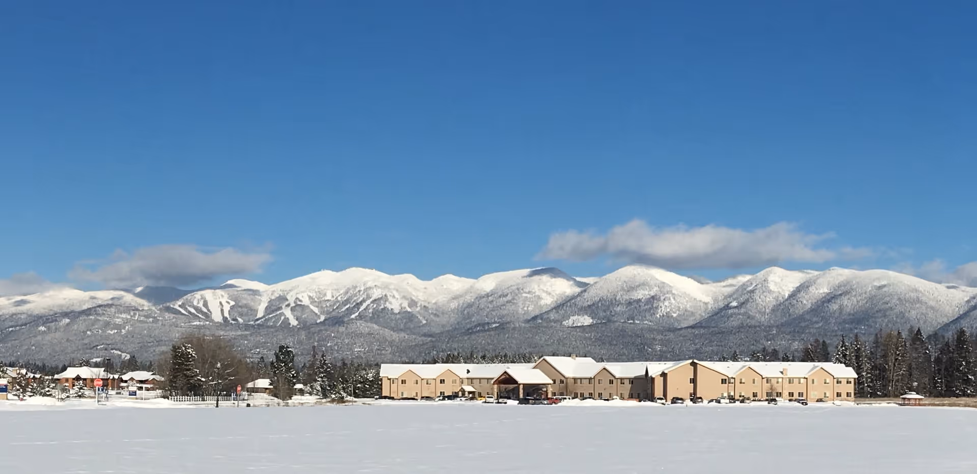 Wide exterior view of The Springs at Whitefish facility with snow-covered ground and parked cars in front, set against a backdrop of snow-capped mountains under a clear blue sky.