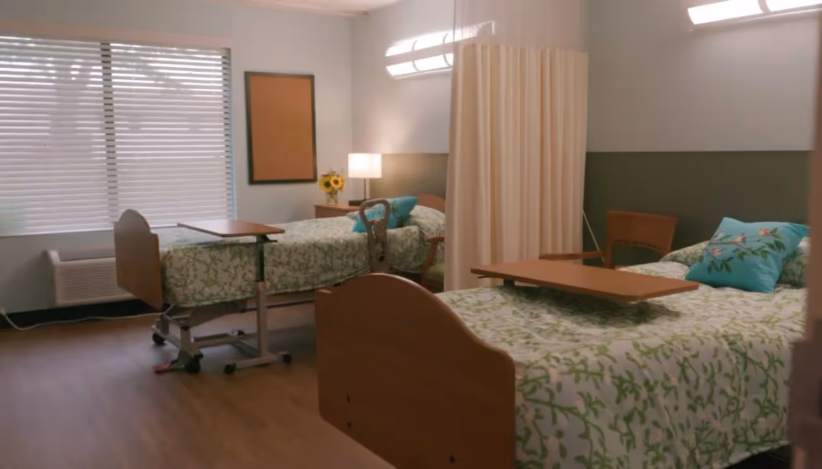 A nursing facility room with two hospital beds, each with green and white patterned bedding and a blue pillow with floral designs. Each bed has an overbed table. There is a privacy curtain between the beds, a wooden chair, a nightstand with a lamp and a vase of sunflowers, and a window with blinds letting in natural light.